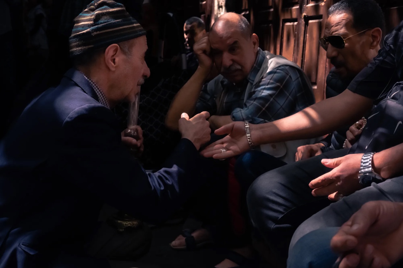 Two older men in traditional caps sitting together, exchanging a firm handshake during a conversation.