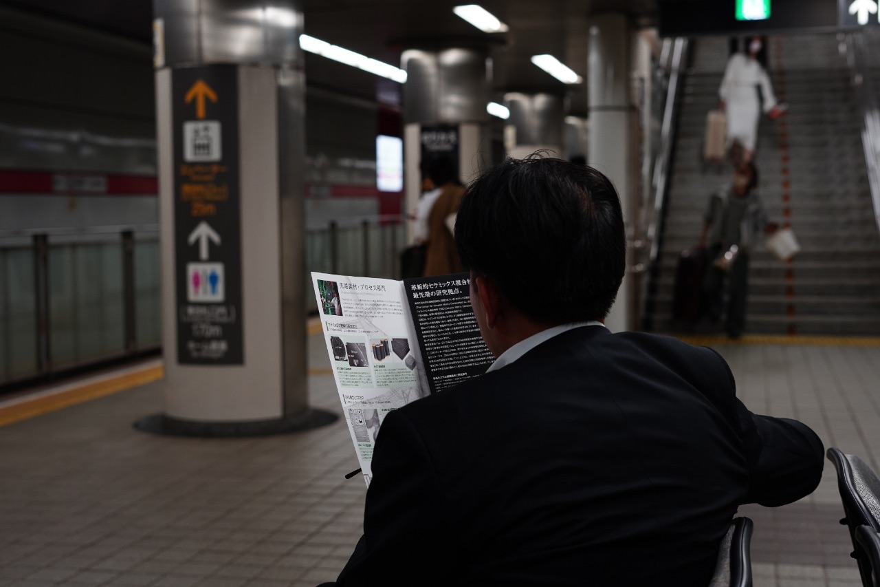 Back-of-head perspective of a man in a dark suit reading a brochure or map inside a busy transit station in Japan