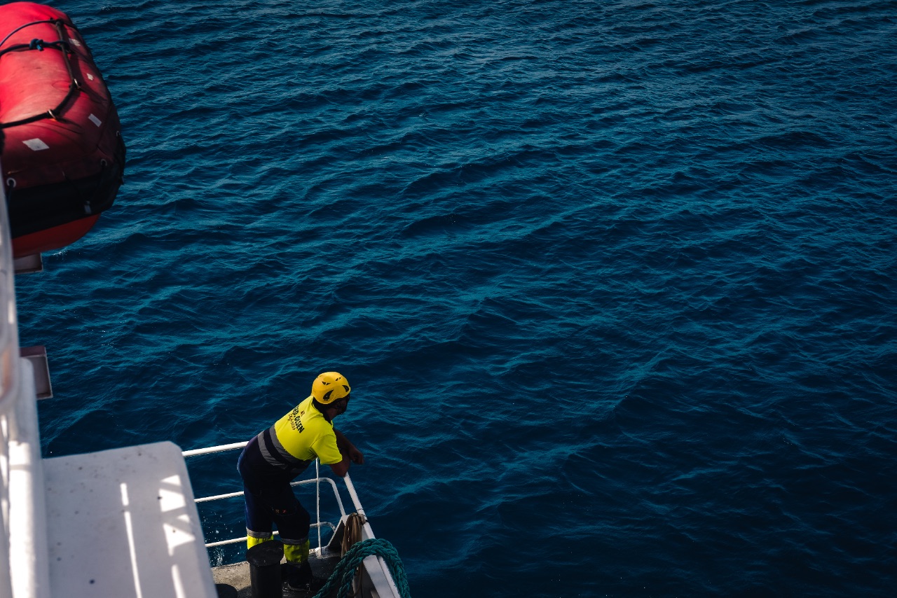 High-angle view of a boat crew member in a yellow high-vis jacket leaning over the stern into deep blue ocean water.